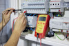 En person använder en röd digital multimeter för att mäta spänning i en elcentral. Displayen visar 397.5 V.