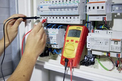 En person använder en röd digital multimeter för att mäta spänning i en elcentral. Displayen visar 397.5 V.