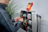 Person testar elektrisk utrustning med en multimeter, med kablar anslutna till en panel nära vägg och växter.