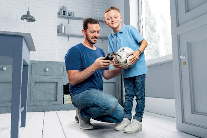 En man och en pojke i blå kläder står inomhus. Mannen håller en kameradrönare; pojken håller en fotboll.