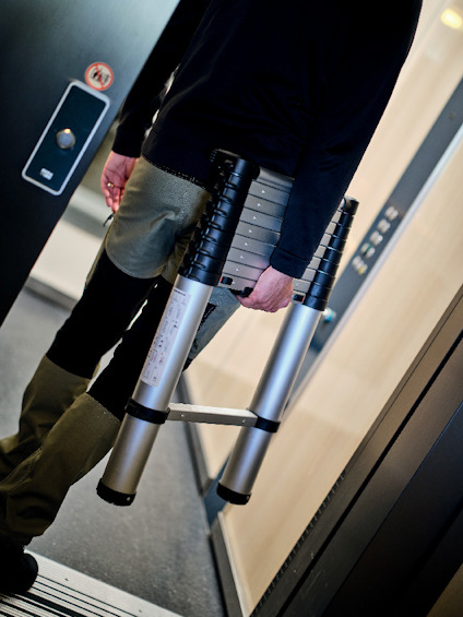 Person bär en hopfällbar stege i en korridor eller entré, klädd i mörka kläder.