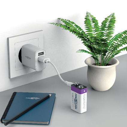 Strömadapter med USB-port ansluten till ett vägguttag, laddar ett 9V-batteri. Anteckningsbok och penna bredvid.