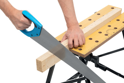 Händer som sågar en träbalk med en blå handsåg placerad på ett arbetsbord med mätmarkeringar.