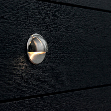 Runt väggljus med metallfinish monterat på svart träpanel.