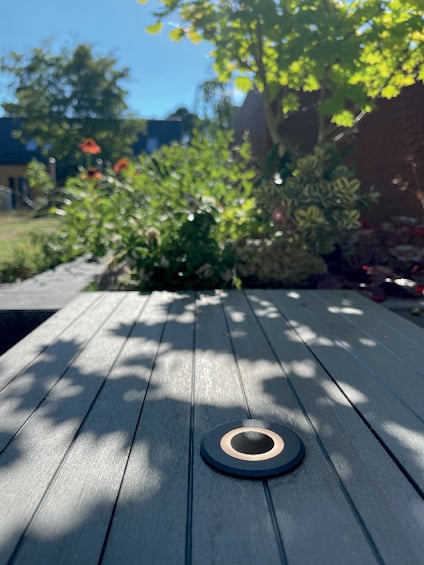 Trädgårdsbord i trä med en infälld, rund belysning på ytan. Skuggor av bladverk syns i förgrunden.