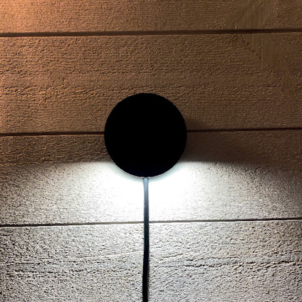 Rund vägglampa med svart hölje lyser upp en ljus träpanelvägg. En kabel hänger ned från lampan.