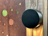 Svart, rund cylinder monterad på träpanel, framför en rostig metallskiva med utstansade cirklar.