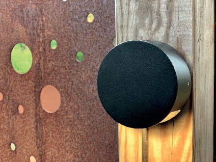 Svart, rund cylinder monterad på träpanel, framför en rostig metallskiva med utstansade cirklar.