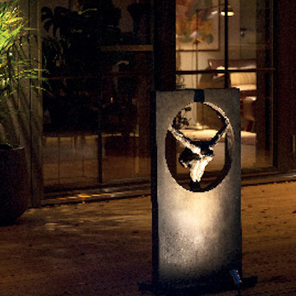 En dekorativ metallskulptur med en cirkulär form och en hjortdesign, placerad på en träveranda omgiven av växter.