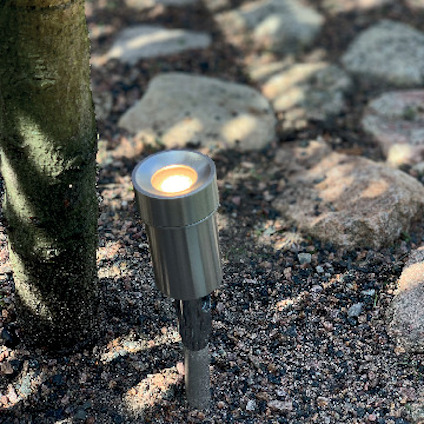 Solcellsdriven marklampa av metall bredvid trädstam och stenar på grus.