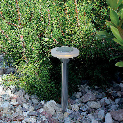 Solcellsdriven trädgårdslampa i metall, placerad bland grus och barrväxter.