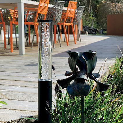 Svart solcellsdriven trädgårdslampa med vattenbubblor, bredvid mörka blommor på en trätrall med orangea stolar i bakgrunden.