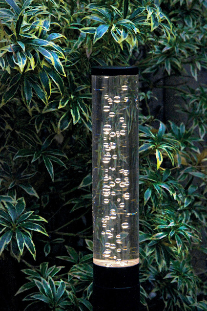 Cylinderformad lampa med luftbubblor inuti, omgiven av gröna blad.