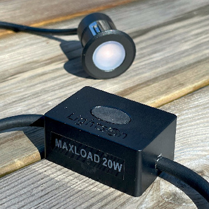 Svart fyrkantig elektronisk komponent med texten "MAXLOAD 20W". Bakgrunden visar en rund lampa på en träyta.