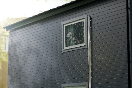 Grå byggnadssida med horisontella paneler och två fönster, belyst av solens strålar.