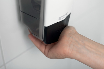 En hand som håller under en automatisk tvålautomat monterad på en vit kakelvägg.