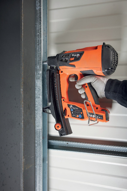 En person håller en orange och svart spikpistol mot en metallram. Personen bär en handske.