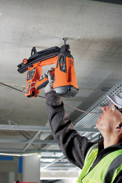 Person i skyddsväst och hjälm använder en orange och svart spikpistol för att fästa något i taket.