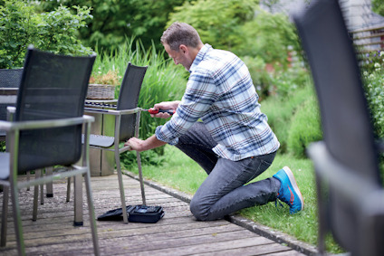 En person i rutig skjorta och jeans placerar ett föremål under en stol på en träaltan utomhus.