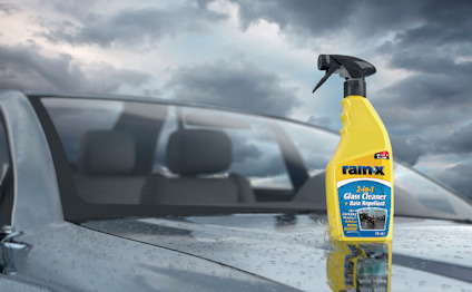 Gul sprayflaska Rain-X 2in1 Glass Cleaner på våt bilhuv, molnig himmel i bakgrunden.