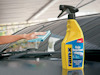 Gul sprayflaska med etikett "Rain Repellent" bredvid en hand som torkar en bilruta med en blå duk.