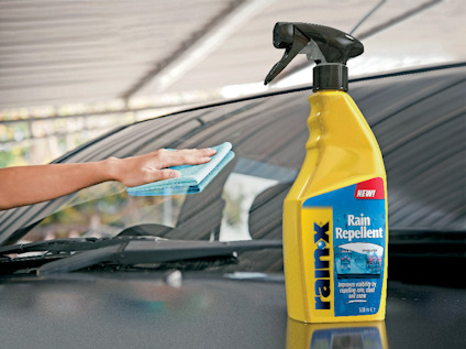 Gul sprayflaska med etikett "Rain Repellent" bredvid en hand som torkar en bilruta med en blå duk.