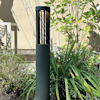 Svart utomhuslampa med cylinderformad skärm placerad bland grön växtlighet vid en vägg.
