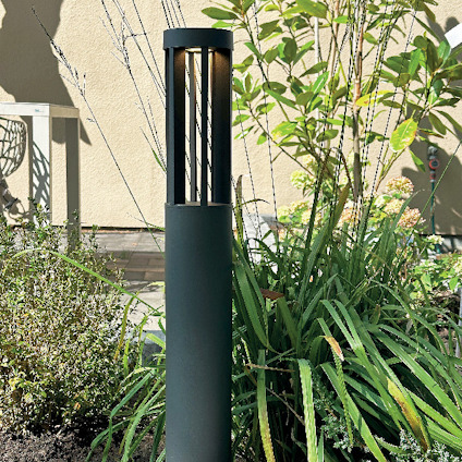 Svart utomhuslampa med cylinderformad skärm placerad bland grön växtlighet vid en vägg.