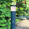 Svart trädgårdslampa med rund, lysande överdel står på en grusgång omgiven av gröna buskar.