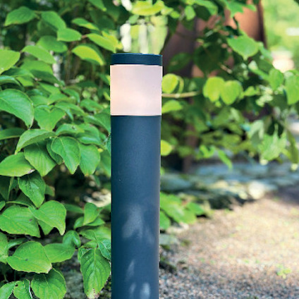 Svart trädgårdslampa med rund, lysande överdel står på en grusgång omgiven av gröna buskar.