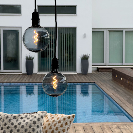 Två glödlampsformade lampor hänger över en pool med trädäck, moderna byggnader i bakgrunden.