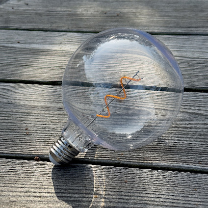 Glödlampa med synlig glödtråd ligger på träplankor i solljus.