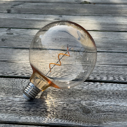 Glödlampa med synlig glödtråd liggande på en träyta utomhus i dagsljus.