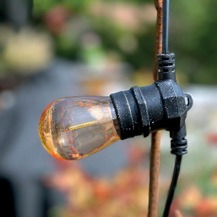 Glödlampa i en svart sockel som hänger vertikalt utomhus mot en suddig bakgrund.