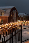 Balkongräcke med julbelysning, snö på marken och rött tegelhus i bakgrunden under skymning.