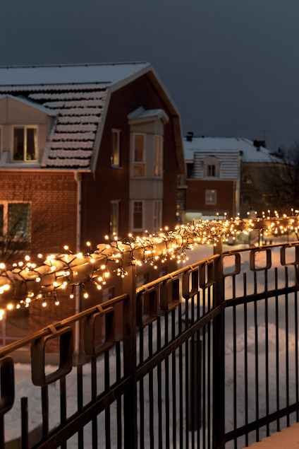 Balkongräcke med julbelysning, snö på marken och rött tegelhus i bakgrunden under skymning.