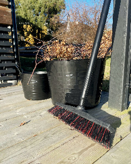Svart sopborste och svarta plastkrukor med kvistar står på en träveranda, träd i bakgrunden.