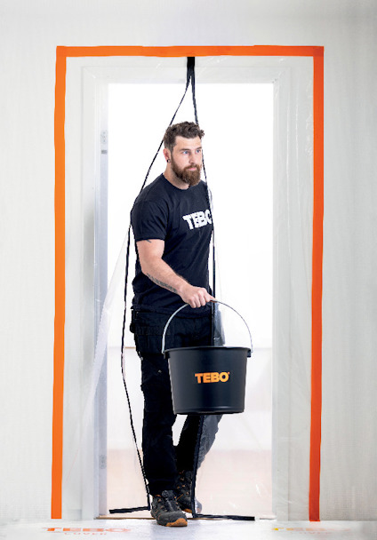 En person med skägg, iklädd Tebo-tröja, går genom en plastskyddad dörröppning och håller en svart hink med Tebo-logga.