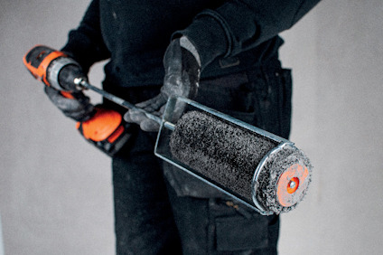 Person håller en borste fäst på en borrmaskin, klädd i svarta arbetskläder och handskar.