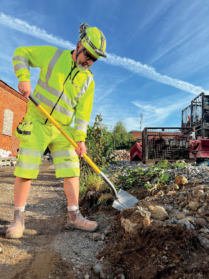 Man i fluorescerande arbetskläder och hjälm använder en spade för att gräva i en stenig jordhög.