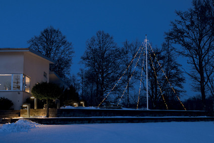 Modernt hus med stora fönster, snötäckt mark och upplysta träd i bakgrunden i skymningen.