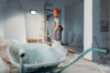 En person i röd hjälm och arbetskläder arbetar i ett renoveringsutrymme med en skottkärra och en stege.