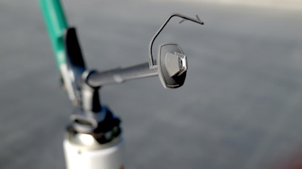 Närbild av en cykeldel med en mindre spegel monterad på styret. Bakgrunden är suddig asfalt.