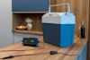 Blå och vit kylbox på bord, bredvid batteriladdare med kablar; frukt och vin syns i bakgrunden.