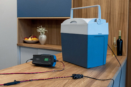 Blå och vit kylbox på bord, bredvid batteriladdare med kablar; frukt och vin syns i bakgrunden.
