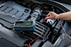 Digital batteriladdare ansluten till bilbatteri i motorutrymme, en hand håller en klämma.