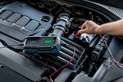 Digital batteriladdare ansluten till bilbatteri i motorutrymme, en hand håller en klämma.