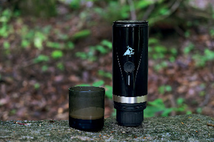 En bärbar kaffemaskin och en kopp står på en stubbe i en skogsmiljö.