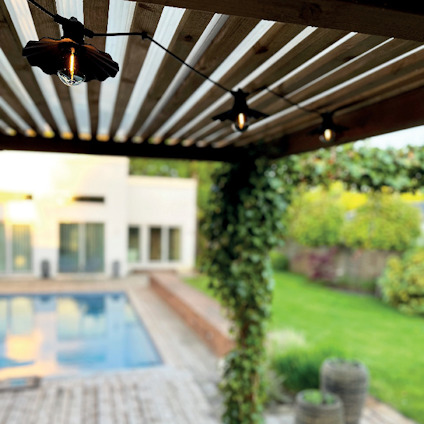 Pergola med hängande lampor, bakom syns en pool, grönska och ett hus i bakgrunden.