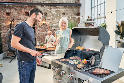 En man grillar kött och grönsaker på en stor gasgrill. En kvinna står bredvid och ler. Två personer sitter vid ett bord i bakgrunden.